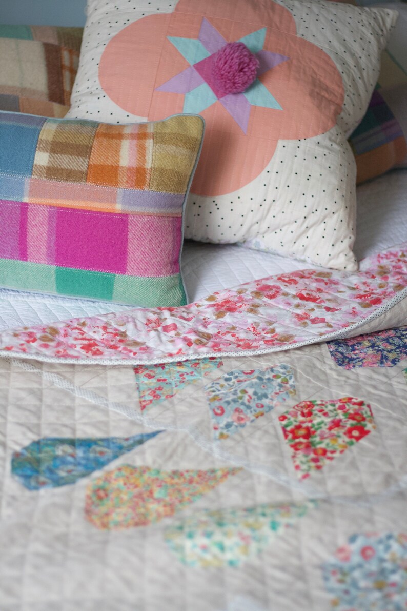 Pu&ograve; includere: Un piumino bianco con un design a patchwork che presenta motivi floreali in tonalit&agrave; di rosa, blu e verde. Il piumino &egrave; ripiegato su un letto con diversi cuscini, tra cui uno con un motivo a pois rosa e bianco e un altro con un motivo a quadri in tonalit&agrave; di rosa, blu e marrone.