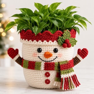 May include: A cream-coloured crochet plant pot cover shaped like a snowman, featuring a red scalloped rim, a green holly appliqué, a striped scarf, two red buttons, and two arms with red mittens, containing a green leafy plant.