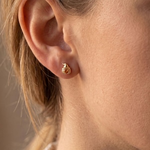 May include: A small gold stud earring shaped like a pumpkin with a witch hat on top. The earring features a polished metallic finish and is secured in the earlobe, showcasing a detailed carved design of a pumpkin with a pointed hat silhouette.