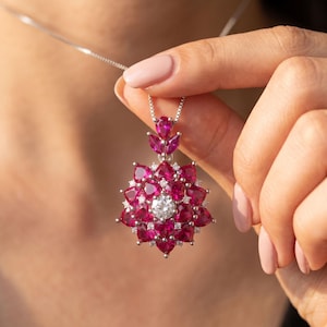 May include: A silver pendant necklace featuring a large, star-shaped cluster of vibrant, deep pink, pear-cut gemstones arranged in multiple layers. A single, clear, round-cut stone sits at the center, surrounded by small, sparkling white accent stones.