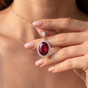 May include: A silver-toned necklace featuring an oval-cut deep red gemstone pendant surrounded by a halo of small, clear, round-cut stones. The pendant is suspended from a delicate silver-toned chain, with additional small stones set into the bail.
