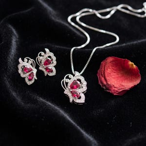 May include: A silver-toned jewelry set featuring a necklace and two stud earrings, each designed in the shape of a butterfly. The wings are accented with small clear stones and feature deep red, pear-shaped stones at the center.
