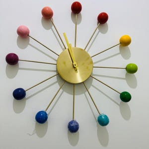 May include: A sunburst wall clock featuring a central gold-toned circular face with matching gold-toned hands. Twelve thin metal spokes extend outward, each tipped with a smooth, spherical ball in a variety of colors including red, yellow, green, blue, purple, and pink.