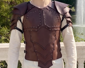 Könnte beinhalten: Ein Brustpanzer aus braunem Leder mit geschichteten Schulterstücken, einer segmentierten Torso-Platte mit sichtbaren Metallnieten und schwarzen Seitengurten, getragen über einem cremefarbenen Langarmshirt und einer braunen Hose.