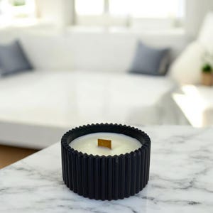 Peut inclure: Une bougie ronde dans un contenant noir cannelé avec une surface de cire blanche et une mèche en bois unique centrée au milieu. L'extérieur du récipient présente une texture nervurée verticale qui parcourt toute la circonférence de l'objet.