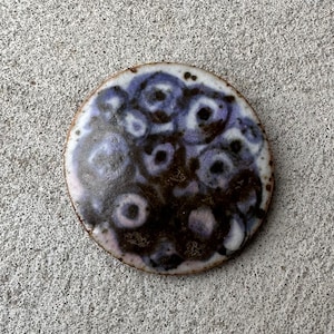 Peut inclure: Un dessous de verre rond en céramique avec un motif abstrait noir et violet. Le dessous de verre a une surface texturée et une finition brillante.
