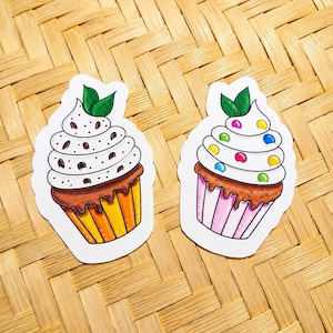 Può includere: Due adesivi fustellati raffiguranti cupcake illustrati. Il cupcake a sinistra ha un pirottino a righe gialle, gocce di cioccolato e due foglie verdi. Il cupcake a destra ha un pirottino a righe rosa, caramelle rotonde colorate e due foglie verdi.