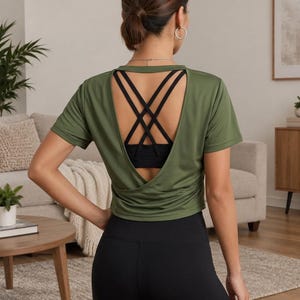 Può includere: Un top sportivo a maniche corte color verde oliva con design a schiena scoperta e dettagli a incrocio. La maglia è indossata sopra un reggiseno sportivo nero e abbinata a leggings neri, per un completo sportivo funzionale e traspirante.