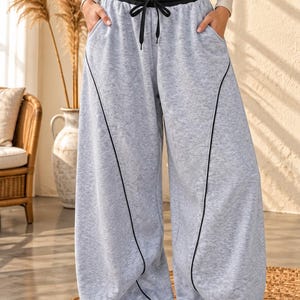 May include: Light gray heathered wide-leg sweatpants featuring a contrasting black elastic waistband with a black drawstring. The pants include side pockets and prominent black piping details that run vertically down the front of each leg.