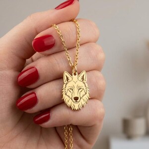 May include: A gold-toned metal necklace featuring a pendant shaped like a wolf head. The pendant displays detailed engraved lines outlining the facial features, ears, and fur of the wolf. The pendant is attached to a delicate gold-toned chain.