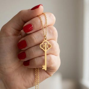 May include: A gold-toned necklace featuring a key-shaped pendant with an ornate, clover-like bow design. The pendant hangs from a delicate, matching gold-toned chain, showcasing a polished finish and a classic, slender key silhouette.