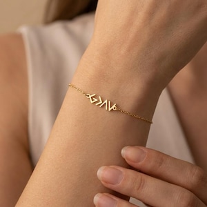 May include: A gold-toned chain bracelet featuring a central metal charm with the text G>AV, which stands for God is greater than the highs and lows. The delicate link chain connects to the charm on both sides, creating a minimalist piece of jewelry.