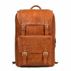 Pode incluir: Uma mochila de couro castanho-claro com uma aba principal grande e um bolso frontal menor, ambos fixados com fivelas de metal em tom de latão. A bolsa inclui uma alça superior e o texto HUB LEATHER CO. em relevo na aba frontal superior.