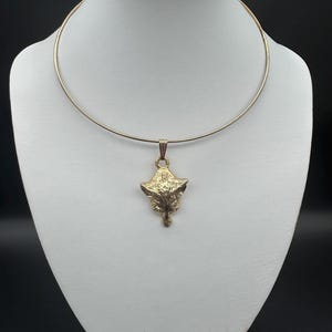 May include: A gold-toned metal collar necklace featuring a textured pendant shaped like a fox head. The pendant is suspended from a thin, rigid circular band that rests around the neck. The metal has a polished finish with detailed relief.