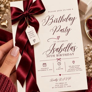Puede incluir: Una tarjeta de invitación rectangular blanca para una fiesta de cumpleaños con un lazo de satén color burdeos en el lado izquierdo. Una pequeña etiqueta blanca unida al lazo dice: THIRTY LOOKS GOOD ON YOU. El texto incluye detalles del 30 cumpleaños.