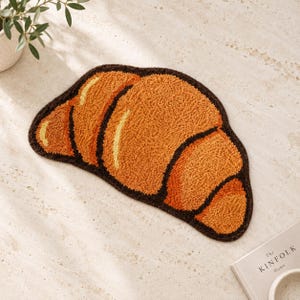 Op de afbeelding: Een getuft tapijt in de vorm van een croissant, met een getextureerd oppervlak in tinten goudbruin, oranje en geel, omlijnd met een donkerbruine rand. Het tapijt heeft een gebogen, gelaagd gebakontwerp met zichtbare getufte garenlussen.