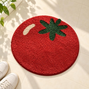 Può includere: Un tappeto rotondo trapuntato a forma di pomodoro. Il tappeto presenta una superficie testurizzata rosso vivace, un dettaglio a stelo verde scuro nella parte superiore e un piccolo segno curvo color bianco sporco sul lato in alto a sinistra.