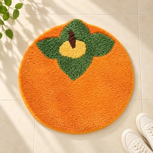 May include: A round, tufted rug shaped like a persimmon fruit. The rug features a large, textured orange circular base with a central design of dark green leaves, a yellow centre, and a small brown stem detail at the very top of the design.
