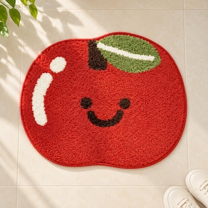 Peut inclure: Un tapis rouge en forme de pomme avec une surface texturée et touffetée. Le motif présente une tige marron foncé, une feuille verte avec un reflet blanc, et un visage souriant avec deux yeux marron foncé et une bouche courbée. Un reflet blanc.