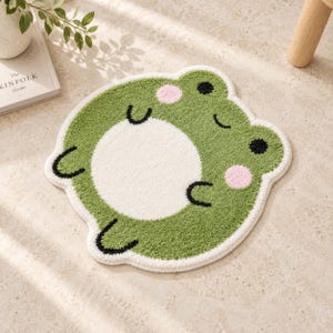 May include: A tufted rug shaped like a frog with a green body, a large white circular belly, two pink circular cheeks, and black eyes and limbs. The rug features a thick white border and a textured, soft pile surface throughout the design.