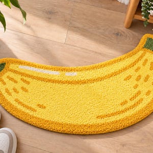 May include: A banana-shaped rug featuring a bright yellow textured surface with darker yellow accents, white highlights along the curve, and dark green tips at both ends. The rug has a tufted pile construction and a curved, elongated silhouette.