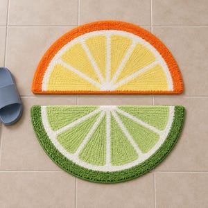 May include: Two semi-circular bath mats shaped like citrus slices. The top mat features a yellow center with a white border and an orange rind. The bottom mat features a light green center with a white border and a dark green rind. A pair of blue slides.