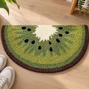 Può includere: Un tappeto trapuntato a semicerchio a forma di fetta di kiwi. Il tappeto presenta un nucleo centrale bianco, polpa verde chiaro, bordi verde scuro e un bordo esterno marrone scuro. Piccole forme nere sono sparse su tutta l'area verde.