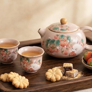 May include: A ceramic tea set featuring a teapot and two cups in a soft pink hue, decorated with painted white rabbits and red strawberries. The set includes a small bowl of fresh strawberries, flower-shaped cookies, and square treats.