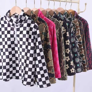May include: A row of long-sleeved quarter-zip pullovers hanging on a rack. The first pullover features a black and white checkerboard pattern. Other pullovers include various prints such as camouflage, solid pink, floral, and lightning bolt patterns.