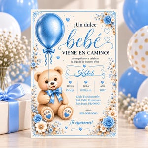 May include: A baby shower invitation card featuring a light brown teddy bear holding a blue balloon, decorated with blue and white flowers. The text reads: Un dulce bebé VIENE EN CAMINO! Kaleb, FECHA 15/sept, HORA 6:00 p.m., AÑO 2027, Club The Butterfly, 245 Calle Primavera, San Juan, PR 00901, RSVP 939-000-0000, Te esperamos!