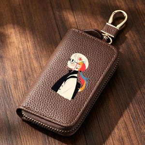 May include: A brown pebbled leather key case with a gold-tone metal swivel clasp and zipper closure. The front features a printed graphic of a stylized anime-style character with blonde hair, glasses, and a colorful ponytail against the dark brown texture.