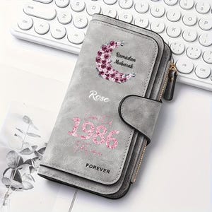 May include: A gray wallet featuring a floral crescent moon graphic with the text Ramadan Mubarak, the name Rose, the year 1986 in pink glitter, and the word FOREVER. The wallet has a snap closure, a side zipper, and a textured matte finish.