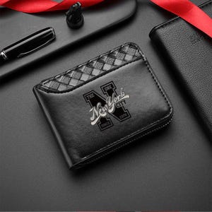 May include: A black bi-fold wallet featuring a woven leather texture on the upper section and a printed graphic on the front. The graphic displays a large letter N with the text New York City, The One, and Since 1978 in white lettering.