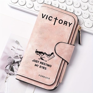 May include: A light pink rectangular wallet with a textured finish, featuring a snap closure and a side zipper. The front is printed with the black text VICTORY, a graphic of a reclining figure, and the text JUST RESTING MY EYES FOREVER.