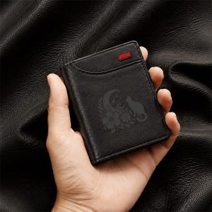 May include: A black leather wallet featuring an engraved design of a cat sitting beside a crescent moon and flowers. The wallet has a small rectangular red accent detail on the upper front edge and visible stitching along the perimeter.