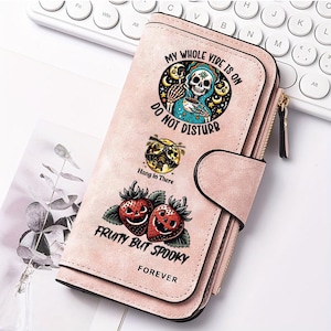 Könnte beinhalten: Ein hellrosa Portemonnaie mit einer Grafik eines kaffeetrinkenden Skeletts und dem Text My Whole Vibe Is On Do Not Disturb, einer Fledermaus mit dem Text Hang in There und zwei Erdbeer-Kürbissen mit dem Text Fruity But Spooky Forever.