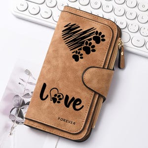 May include: A tan, long wallet with a textured finish featuring a black graphic of a heart made of lines and paw prints. The word LOVE is printed in black with a paw print inside the letter O, and the word FOREVER is printed below it in black.