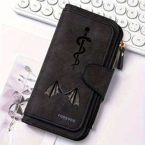 May include: A black rectangular wallet featuring a vertical zip closure, a snap button flap, and a matte finish. The front displays a black graphic of a staff with a coiled snake, a pair of bat wings, and the word FOREVER printed in white.
