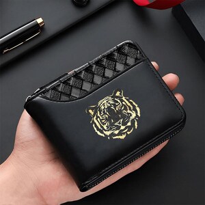 May include: A black bi-fold wallet featuring a gold tiger head graphic on the front. The upper portion of the wallet has a black woven leather texture, while the lower section is smooth black leather. It includes a silver-toned metal zipper closure.