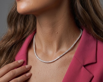 May include: A silver-toned tennis necklace featuring a continuous row of small, round, clear, faceted stones set in a metallic chain. The necklace rests against the neck, showcasing a uniform design with a bright, reflective finish throughout.