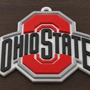 May include: A three-dimensional Ohio State University logo ornament featuring a central red block O with a black letter O inside. The word OHIOSTATE is written in bold black capital letters across the center. The design has white and gray borders.