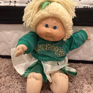 May include: A Cabbage Patch Kids doll with light skin, blue eyes, and blonde yarn hair tied with a green ribbon. The doll wears a green long-sleeved sweatshirt with the Cabbage Patch Kids logo in white text and a green and white pleated skirt.