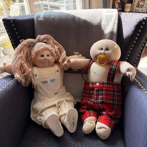May include: Two fabric dolls with soft bodies and yarn hair. The left doll has long pinkish-brown hair, a white overall outfit, and a signature on the left foot. The right doll is bald, has a pacifier in its mouth, and wears red plaid overalls.