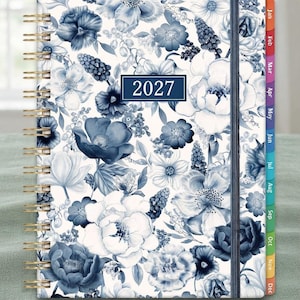 May include: A 2027 planner featuring a white cover with a dense blue floral pattern, gold-tone spiral binding, and a dark blue elastic closure. The side edge displays twelve colorful monthly tabs labeled from Jan to Dec in white text.