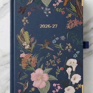 May include: A navy blue hardbound planner with the text 2026-27 printed in gold. The cover features a colorful botanical illustration of hummingbirds, pink hibiscus, white calla lilies, and various green leaves. It has gold-toned metal corner protectors.