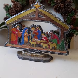 May include: A decorative nativity scene ornament featuring a stable structure with a yellow star at the peak. The scene depicts figures in colorful robes, a manger, and animals. The base includes the text FELIZ NATAL in raised lettering.