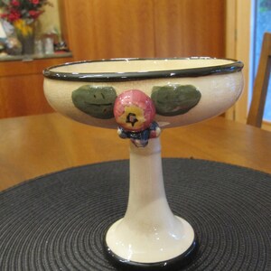 May include: A ceramic pedestal bowl with a wide, shallow cream-colored bowl and a tall, flared base. The bowl is decorated with green leaf shapes and a pink and red fruit-like figure. The rim and base are trimmed with black.