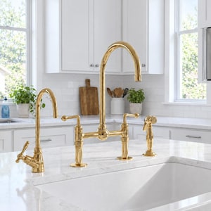 Può includere: Un set di rubinetti da cucina in oro lucido composto da un rubinetto a ponte ad arco alto con due maniglie a leva, un rubinetto laterale più piccolo e una doccetta abbinata. Gli elementi hanno una finitura metallica su un piano in marmo.