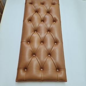 May include: A rectangular bench cushion upholstered in smooth tan leather featuring a deep diamond tufted pattern with matching fabric-covered buttons. The cushion has a structured piped edge and a consistent, uniform surface texture throughout.