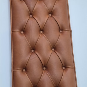 Può includere: Un pannello rettangolare in pelle color cuoio con un design trapuntato caratterizzato da bottoni distanziati uniformemente che creano un motivo a rombi sulla superficie. Il pannello ha una finitura liscia e un bordo profilato e strutturato.
